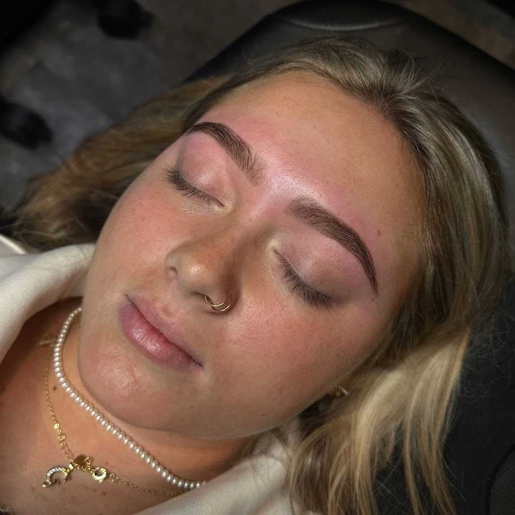 Woman with freshly shaped eyebrows, eyes closed, wearing layered necklaces and a nose ring.