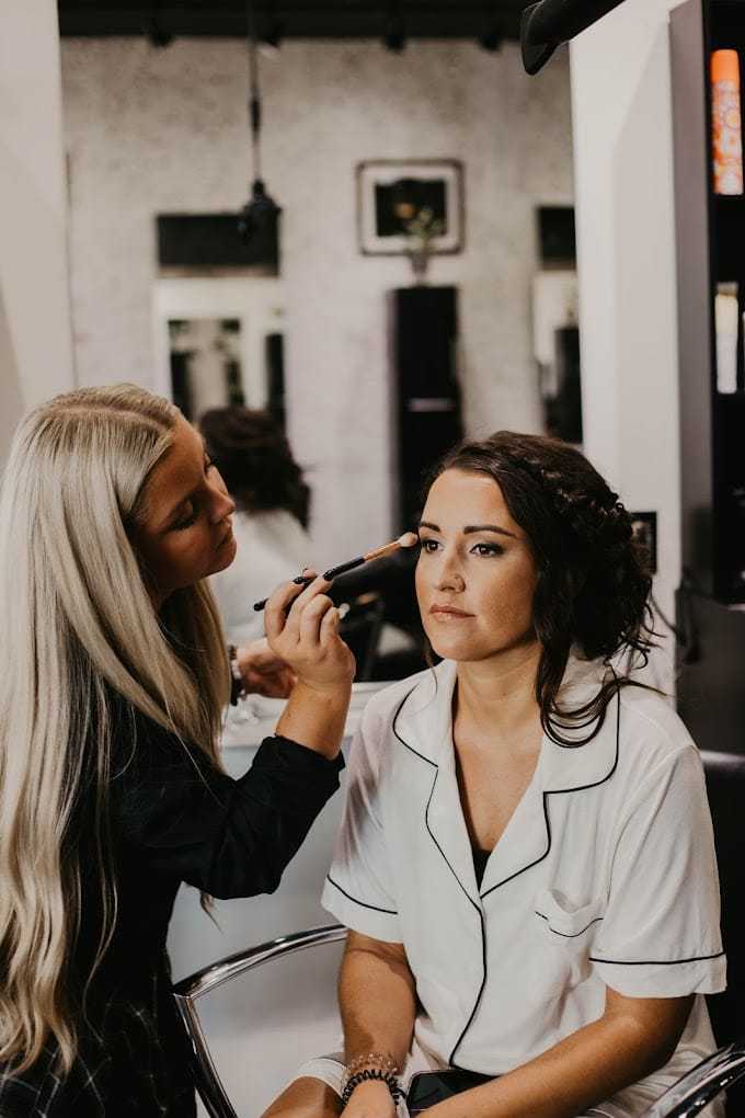 Hair stylist applying makeup to a seated woman in a salon.