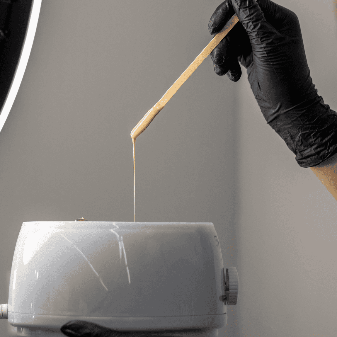 Hot wax on spatula over a waxing pot, held by a gloved hand, in a salon setting.